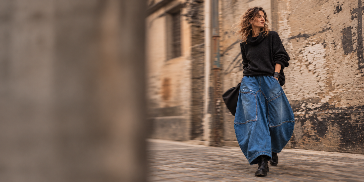 Vrouw in een opvallende blauwe ballonjeans en zwarte oversized trui loopt door een karaktervolle straat. Editorial modebeeld van LEEZZA Fashion.